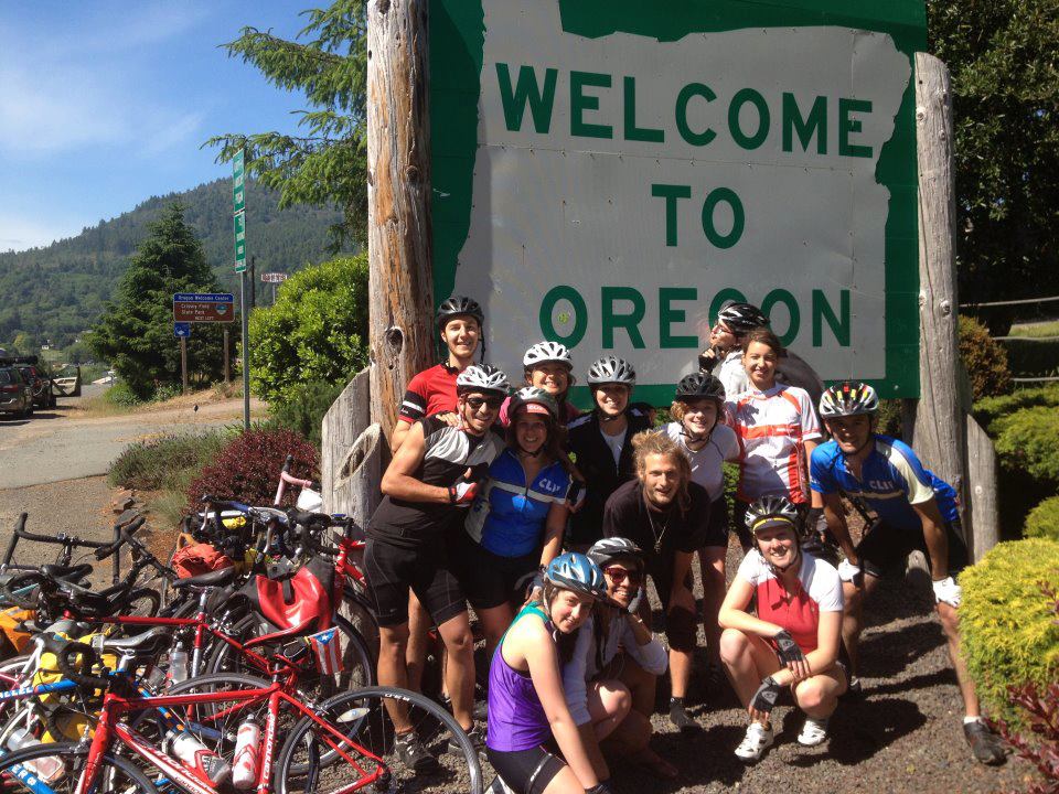 Co-cycle members on bikes, standing next to "Welcome to Oregon Sign"