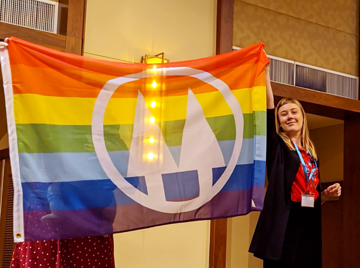 Image of a smiling person holding up the Co-op Flag, which has a rainbow background with the twin pines logo in the middle in white