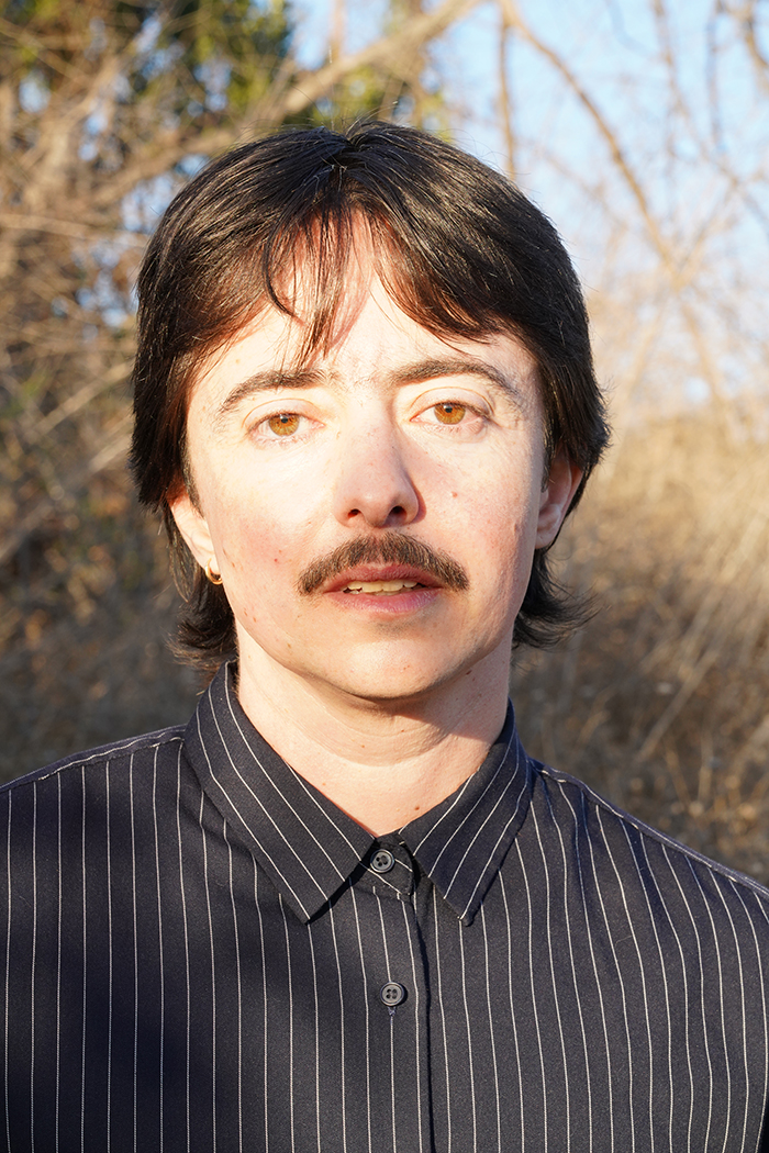 Head shot of a white man with brown hair and mustache, standing outside. He is wearing a black and white button-up shirt. 